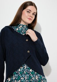 Cardigan blu navy con un motivo a foglia testurizzato, dotato di un colletto a punta e due grandi bottoni, indossato sopra un vestito a fantasia.