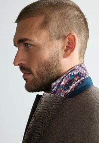 Brown herringbone blazer with a blue collar and a floral patterned inner collar in shades of pink, blue, and white. Short, closely cropped hair.