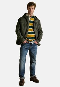 Tamsiai žalia striukė su gobtuvu, su dviem krūtinės kišenėmis, dėvima ant daugiaraugių juostuotų polo marškinėlių, ir derinama su mėlynomis džinsomis ir rudais batais.