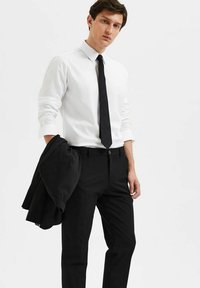 Witte dress shirt, zwarte stropdas, zwarte pantalon, en blazer over de arm. Eenvoudig ontwerp, gladde textuur en getailleerde silhouette.