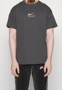 Šedé bavlněné tričko s malým žlutým logem Nike a nápisem na hrudi. Kulatý výstřih a krátké rukávy, doplněné černými tepláky.