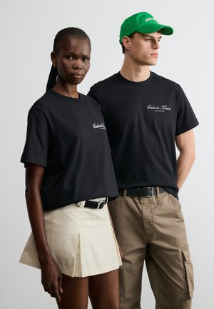 Két ember, fekete Calvin Klein pólót viselve; egy nő bézs szoknyában és egy férfi khaki nadrágban és zöld sapkában, egyszerű háttér előtt állnak.