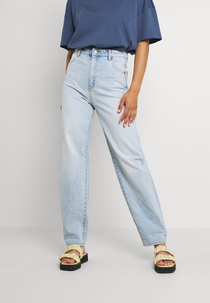 Jeans de cintura alta azuis claros com pernas retas, tecido de denim, ligeira descoloração. Usados com sandálias amarelas com folhos e uma blusa azul solta.