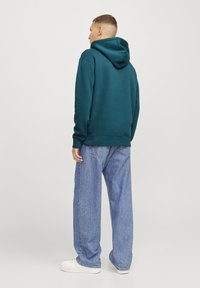 Teal hettegenser med en frontlomme og hette, kombinert med lyseblå vide jeans og hvite sneakers, sett fra baksiden.