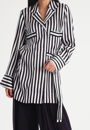 Femme portant un blazer à rayures verticales noires et blanches avec de larges poignets et un pantalon noir à jambes larges.