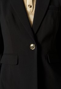 Blazer nero su misura con una texture liscia, caratterizzato da un bottone dorato e due tasche frontali. Sotto, un top lavorato a maglia beige con chiusura a bottone.