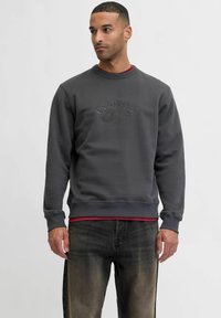 Szary sweter z okrągłym dekoltem, z wytłoczonym napisem „ORIGINALS”, zestawiony z bordową koszulą pod spodem oraz ciemnymi dżinsami.