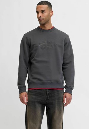 Szary sweter z okrągłym dekoltem, z wytłoczonym napisem „ORIGINALS”, zestawiony z bordową koszulą pod spodem oraz ciemnymi dżinsami.