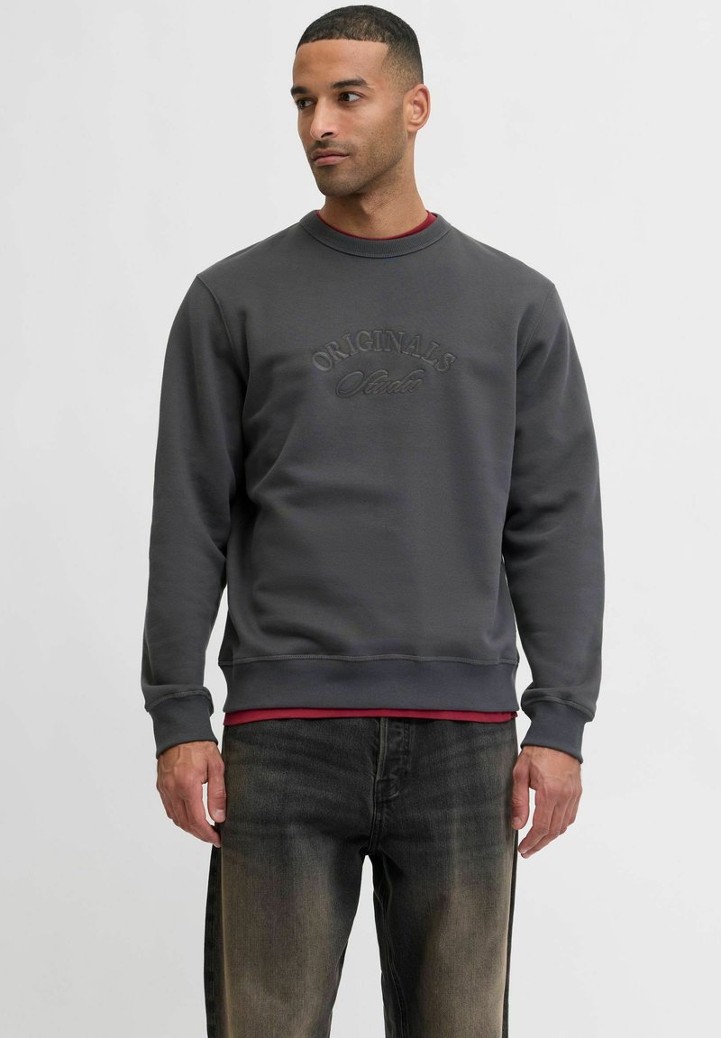 Szary sweter z okrągłym dekoltem, z wytłoczonym napisem „ORIGINALS”, zestawiony z bordową koszulą pod spodem oraz ciemnymi dżinsami.