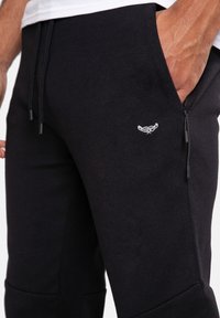 Gros plan sur une personne portant un pantalon de jogging noir avec des poches latérales zippées et un petit logo blanc brodé près de la poche.