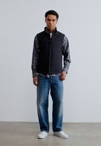 Άνδρας που φοράει μαύρο αμάνικο γιλέκο πάνω από πουκάμισο με καρό σχέδιο, μπλε τζιν και λευκά αθλητικά παπούτσια, στέκεται μπροστά σε έναν απλό φωτεινό τοίχο.