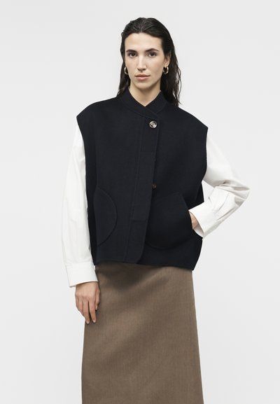 Γυναίκα με σκούρα μαλλιά φορεί χρυσούς κρίκους στα αυτιά, ένα μαύρο αμάνικο γιλέκο με κουμπιά πάνω από λευκό πουκάμισο με μακριά μανίκια και καφέ φούστα.