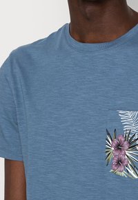 Modré bavlněné tričko s krátkým rukávem, které má náprsní kapsu zdobenou tropickými květinovými vzory v růžové, zelené a bílé barvě.