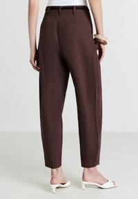 Donna che indossa pantaloni scuri a vita alta e a sigaretta marrone scuro, sandali con tacco bianchi, una maglietta bianca e due braccialetti rigidi al polso sinistro.
