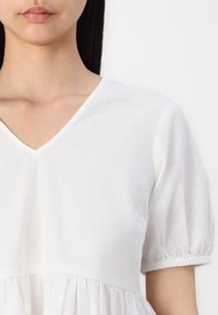 Fehér pamut felső V-nyakkal, rövid puff ujjakkal és ráncolt derékkal, puha tapintású és könnyű anyagból készült.