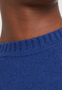 Sweter dziany z prążkowanym okrągłym dekoltem, wykonany z niebieskiej wełny, z teksturowaną powierzchnią. Zbliżenie pokazuje detal dekoltu i tkaniny swetra.