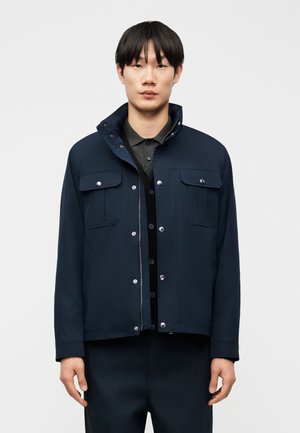 Mann mit schwarzen Haaren, der eine marineblaue Jacke über einem dunkelgrauen Poloshirt trägt, vor einem schlichten hellgrauen Hintergrund.