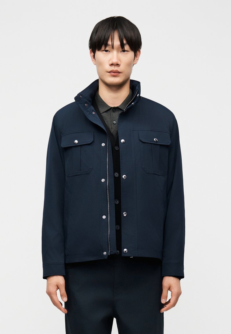 Άνδρας με μαύρα μαλλιά που φοράει σκούρο μπλε μπουφάν πάνω από ένα σκούρο γκρι πόλο πουκάμισο, με φόντο απλό ανοιχτό γκρι.