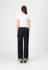 Jeans largas de denim azul escuro com bainha dobrada, combinados com uma t-shirt justa branca, vistos por trás. Design minimalista e linhas limpas.