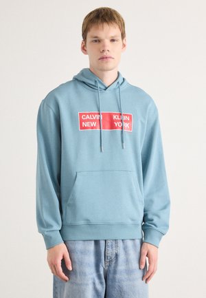 Light blue hoodie made of cotton with a front kangaroo pocket. Features a red rectangular logo with white text reading "CALVIN KLEIN NEW YORK."