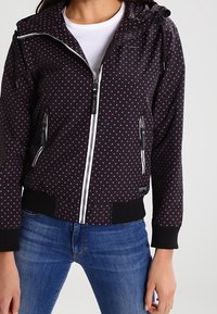 Černá bunda s červenými puntíky, přední zip, dvě kapsy na zip a kapuce. Má žebrované manžety a lem pro těsné uchycení.