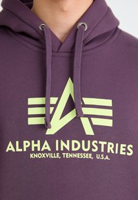 Hoodie violet avec un motif frontal en vert clair représentant un emblème "A" et le texte "ALPHA INDUSTRIES, KNOXVILLE, TENNESSEE, U.S.A."