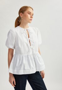 Blouse en coton blanc avec des manches ballon, décolleté en trou de clé et liens à l'avant ; elle présente un ourlet peplum et une texture lisse. Portée avec un jean foncé.