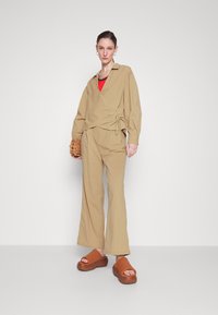 Beige Baumwoll-Wickeloberteil mit langen Ärmeln, kombiniert mit passender, lockerer Hose. Enthält ein rotes Unterhemd und braune Plateausandalen.