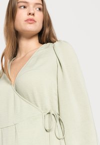 Blusa envolvente verde-clara com um decote profundo em V, mangas franzidas e cintura atada. Composta por um tecido leve e texturizado.