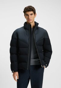 Donkerblauwe pufferjack met een hoge kraag, contrasterende schouderpanelen en een ritssluiting aan de voorkant. Voorzien van knoopsluiten bij de mouwen en een soepele textuur.