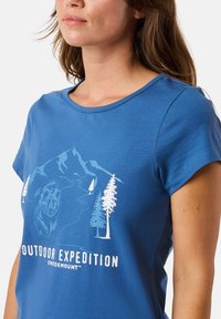 Blå bomulls t-shirt med korta ärmar som har en vit grafisk design av en björn, berg och träd, med texten "OUTDOOR EXPEDITION."