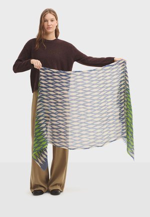Écharpe légère présentant un motif géométrique dans des tons de bleu, blanc et vert. Texture douce avec des bords à franges, tenue par un modèle.