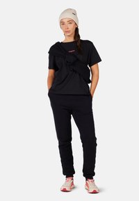 Černé tričko s logem, v kombinaci s černými tepláky a atletickými botami. Model má na hlavě pletenou béžovou čepici a černý svetr přehozený přes ramena.