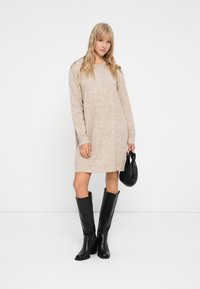 Μπεζ πλεκτό φόρεμα με μακριά μανίκια, στρογγυλή λαιμόκοψη και άνετη εφαρμογή, συνδυασμένο με μαύρες κολάν μπότες μέχρι το γόνατο και μια μικρή μαύρη τσάντα.