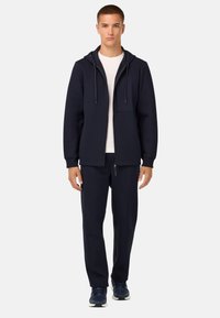 Mornarsko modra zip-up dukserica s kapuco, ki vključuje sprednji žep in vrvice, v kombinaciji z usklajenimi trenirko hlačami in športnimi čevlji.