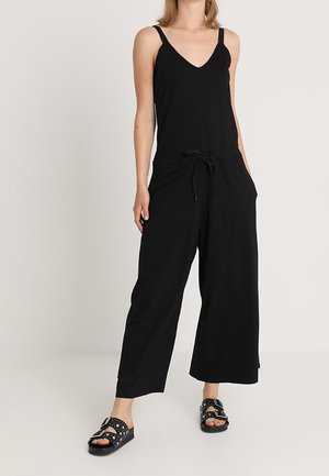 Combinaison noire avec un décolleté en V et des bretelles fines, dotée d'une taille à cordons et de jambes larges. Portée avec des sandales noires ornées de clous en argent.