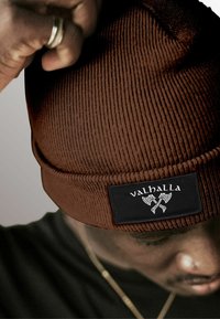 Brown ribbed beanie with a black patch featuring the text "valhalla" and ax design. Warm material with a folded cuff at the base.