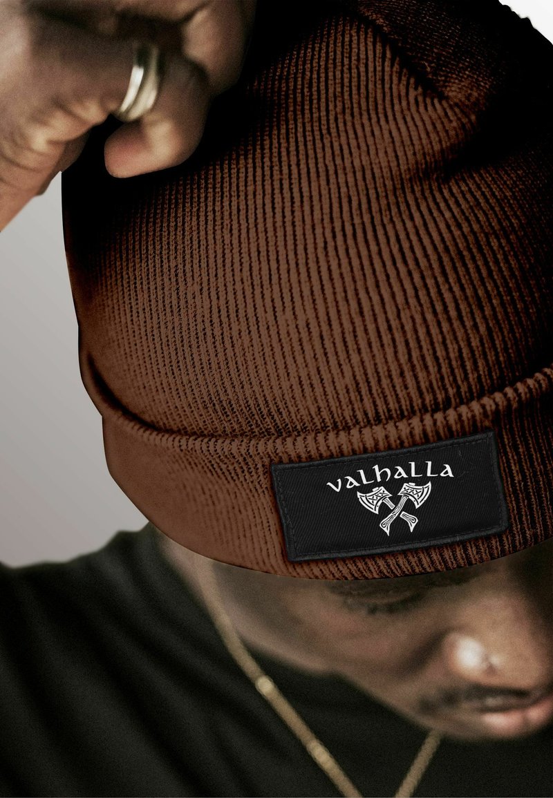 Brown ribbed beanie with a black patch featuring the text "valhalla" and ax design. Warm material with a folded cuff at the base.