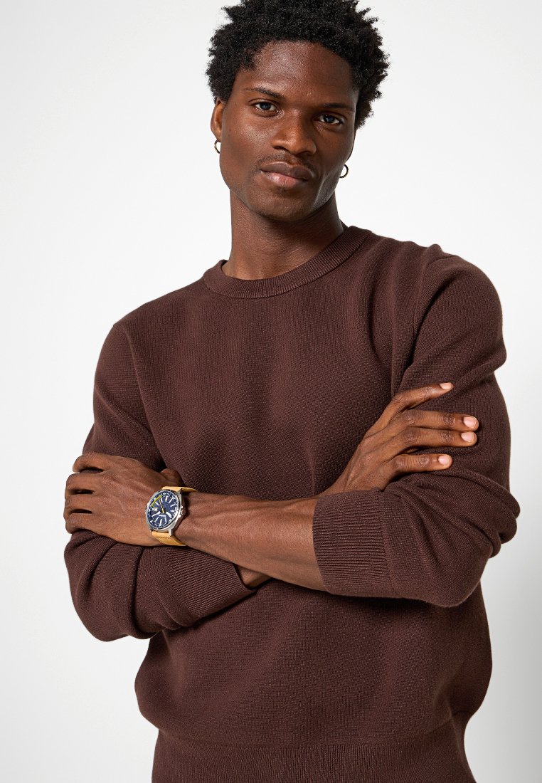 Man met donker krullend haar en kleine ringoorbellen, gekleed in een bruine trui en een horloge, met zijn armen gekruist, tegen een effen witte achtergrond.