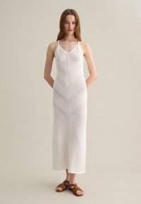 Maxi dress bianco lavorato a maglia con spalline sottili, featuring un design geometrico a openwork e un corpino aderente. Indossato con sandali marroni con lacci.