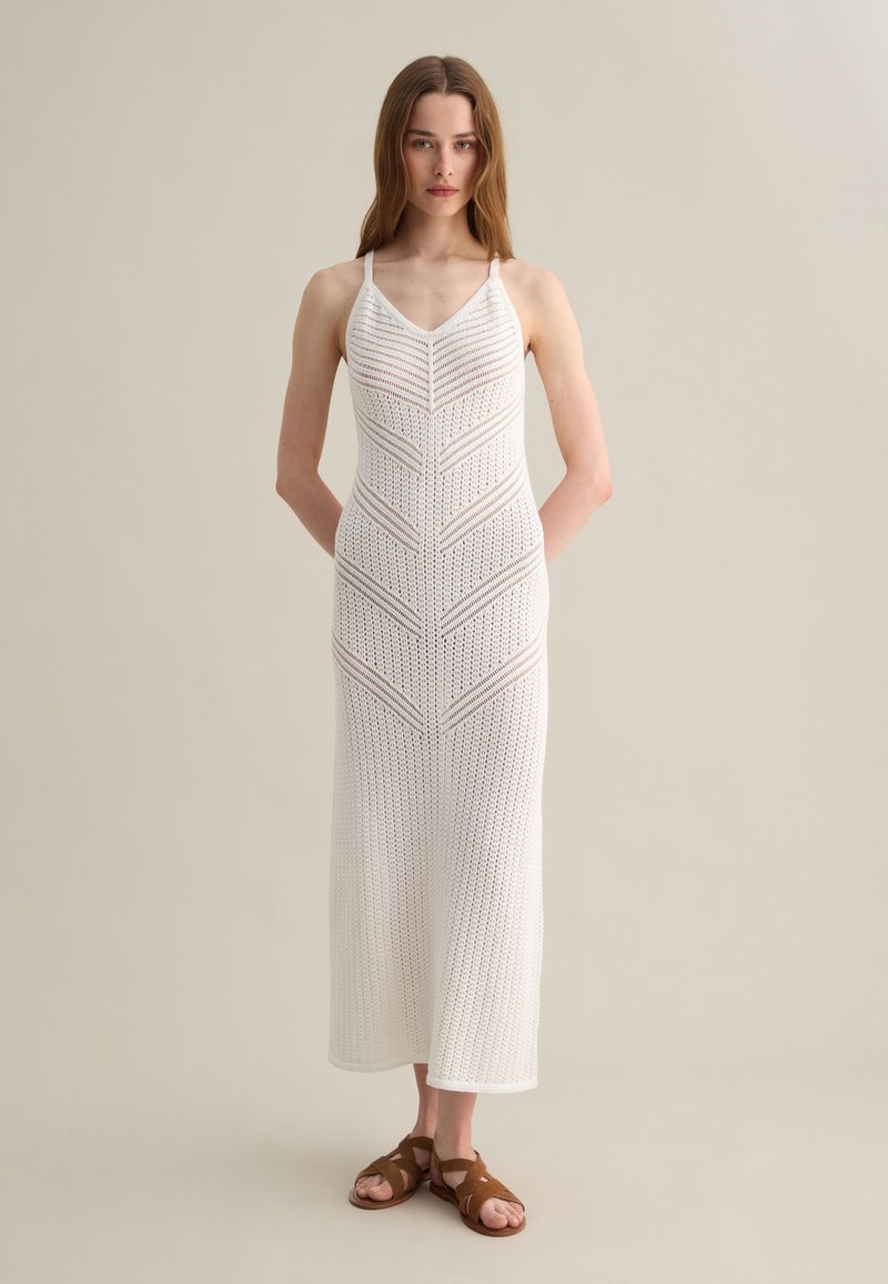 Maxi dress bianco lavorato a maglia con spalline sottili, featuring un design geometrico a openwork e un corpino aderente. Indossato con sandali marroni con lacci.