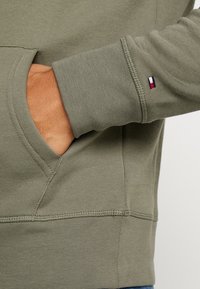Un sweat à capuche vert olive avec une poche kangourou, des poignets côtelés et un petit logo multicolore sur la manche. Tissu doux et texturé.