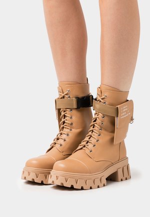 Tan lace-up combat boots with a chunky sole, featuring a fabric strap and pouch on the ankle. Smooth leather texture and silver hardware accents.