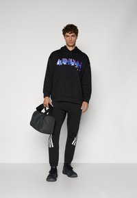 Feliz de capuz preto com gráfico multicolorido "ARIDA", combinado com calças de moletom pretas com riscas brancas. Saco de desporto preto, calçado casual.