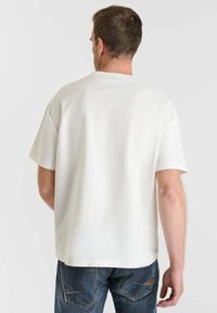 Uomo di spalle che indossa una semplice maglietta bianca a maniche corte e jeans blu su uno sfondo chiaro e uniforme.