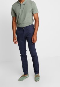 Polo shirt in cotone verde, pantaloni in cotone blu navy e scarpe slip-on in cotone verde chiaro con lacci. Outfit casual con design e colori semplici.