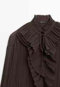 Blouse marron foncé en tissu plissé, dotée d'un col haut, d'accents volants et d'un détail à nouer sur le devant. Texture lisse.