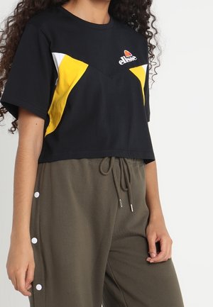 Osoba ubrana w czarny krótki top z żółto-białym wzorem oraz brązowe spodnie z troczkami i białymi guzikami po boku.