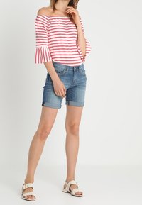 Top de rayas rojas y blancas con hombros descubiertos y mangas acampanadas, combinado con pantalones cortos de mezclilla azul claro y sandalias blancas.