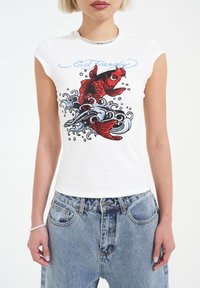 White fitted t-shirt with cap sleeves, featuring vibrant red fish and wave designs in contrasting colors. Fabric appears smooth with a slight sheen.